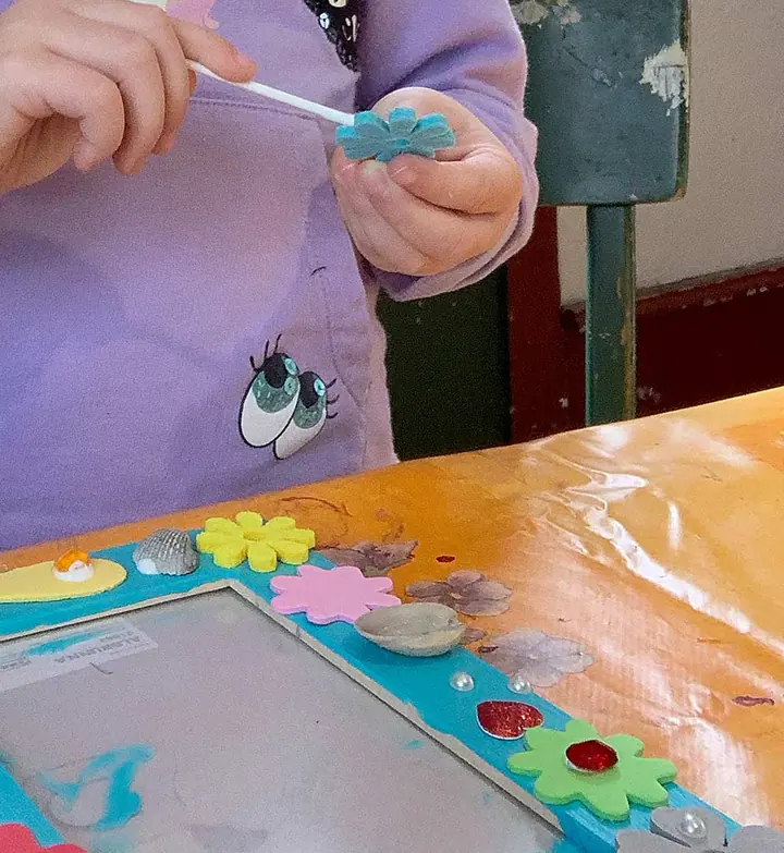 Kinderhände kleben eine Schaumstoffblume auf einen türkisen Bilderrahmen